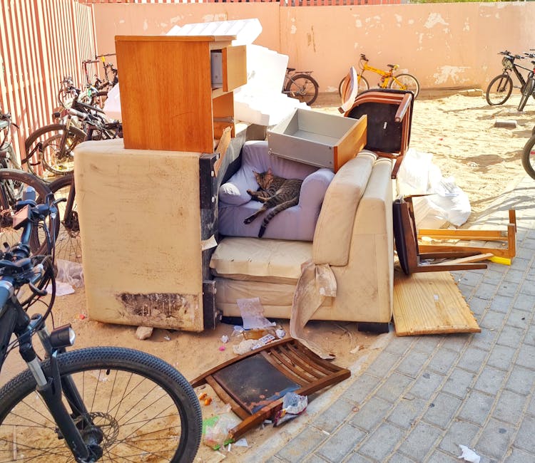 A pile of discarded household furniture and waste materials outdoors on a paved area, including an old, stained sofa with a visible patch of dirt at the bottom, a small purple armchair with a tabby ca