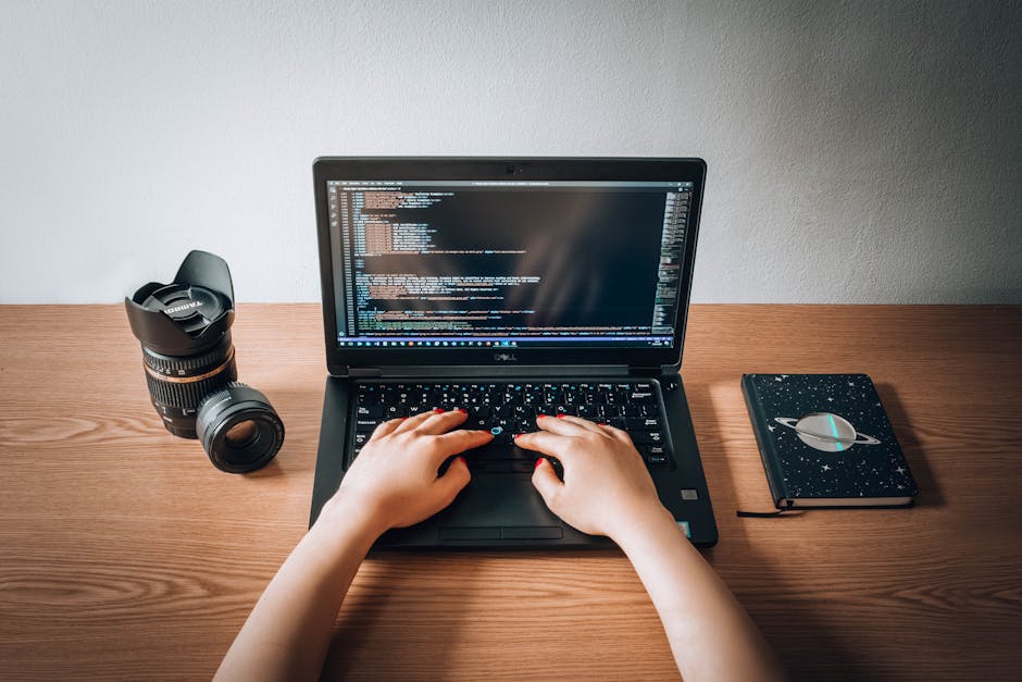 A person typing on a black laptop keyboard placed on a wooden desk, with the laptop screen displaying lines of computer code. To the left of the laptop, there is a black camera with a zoom lens, and t