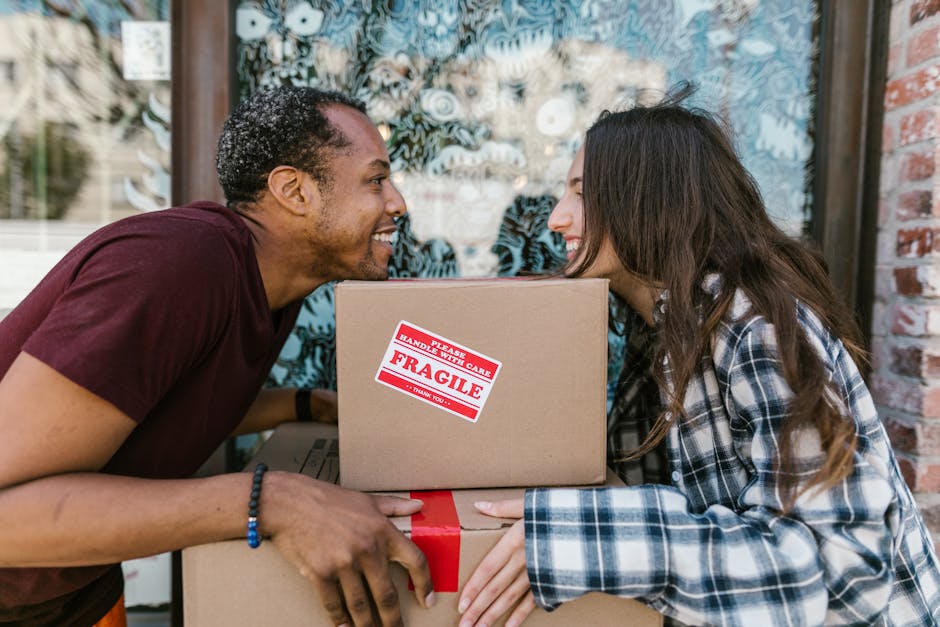 Two individuals engaged in a home relocation process outdoors in front of a building with large windows and brick walls. The man, with short, curly hair and wearing a maroon t-shirt, is smiling and le