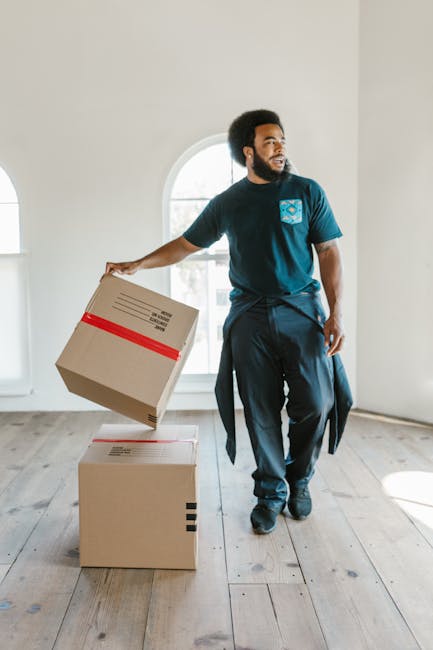 Two individuals engaged in a home relocation process outdoors in front of a building with large windows and brick walls. The man, with short, curly hair and wearing a maroon t-shirt, is smiling and le
