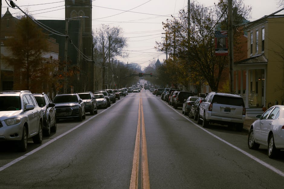 A straight residential street in Merton with parked cars lining both sides of the road, including sedans, SUVs, and hatchbacks in various colours such as white, silver, and black. The street surface i