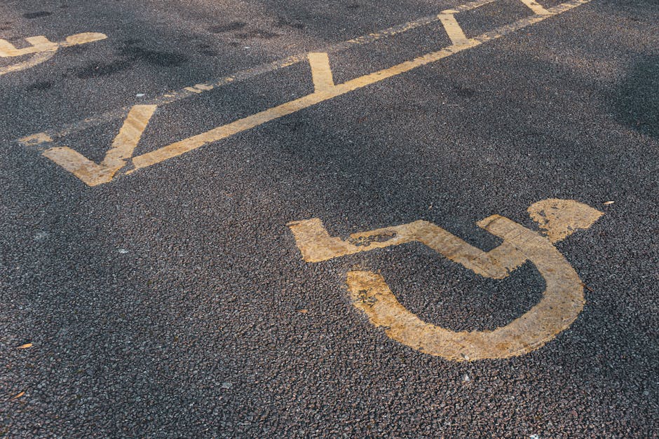 Close-up of a designated accessible parking space on an asphalt surface, featuring a yellow wheelchair symbol painted on the ground with some signs of wear and peeling. The space includes white diagon