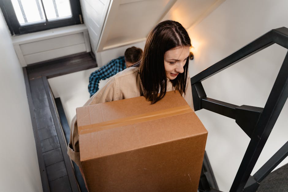 A woman with dark brown hair, wearing a beige coat, is carrying a large rectangular cardboard box up a narrow staircase in a residential property. Behind her, a man wearing a blue and black checked sh