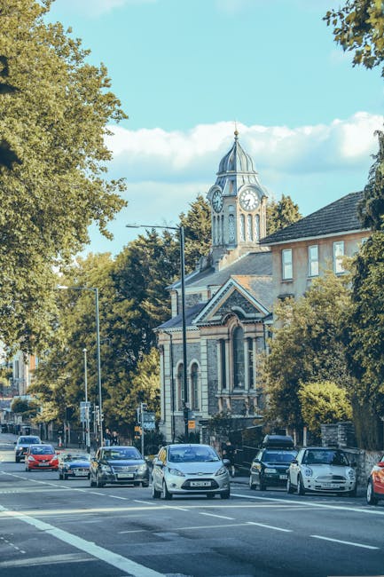A wide view of a street in SW19 showing several parked cars along the curb, with some in motion, on a sunny day with a clear blue sky. Prominent in the background is a historic building with a clock t